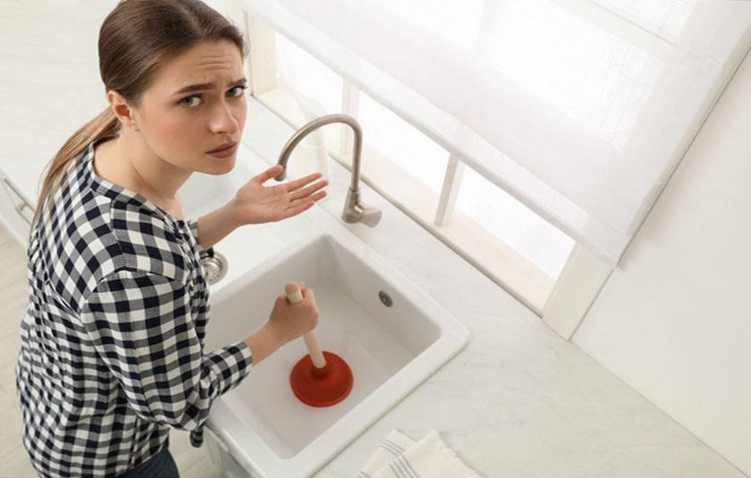 A surprised woman is holding a plunger while trying to unclog the kitchen sink.